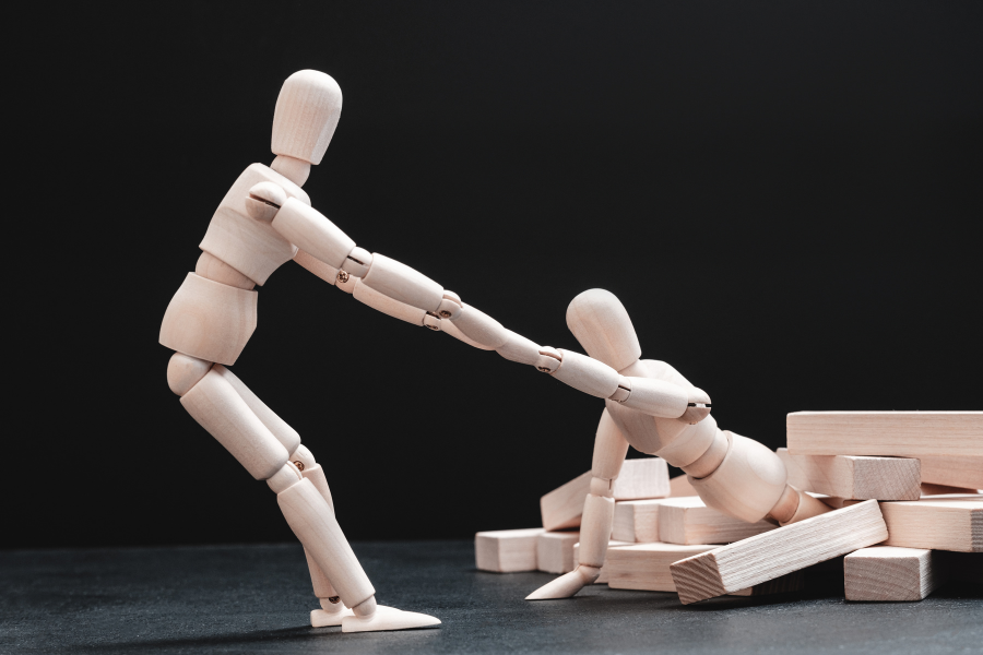 Деревянная фигурка помогает другой выбраться из беспорядка — образ поддержки, восстановления и человеческой устойчивости.